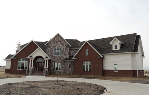 A large brick and stone house with multiple gables, a mix of brick and white siding, and a partially finished driveway. The yard appears to be under construction.