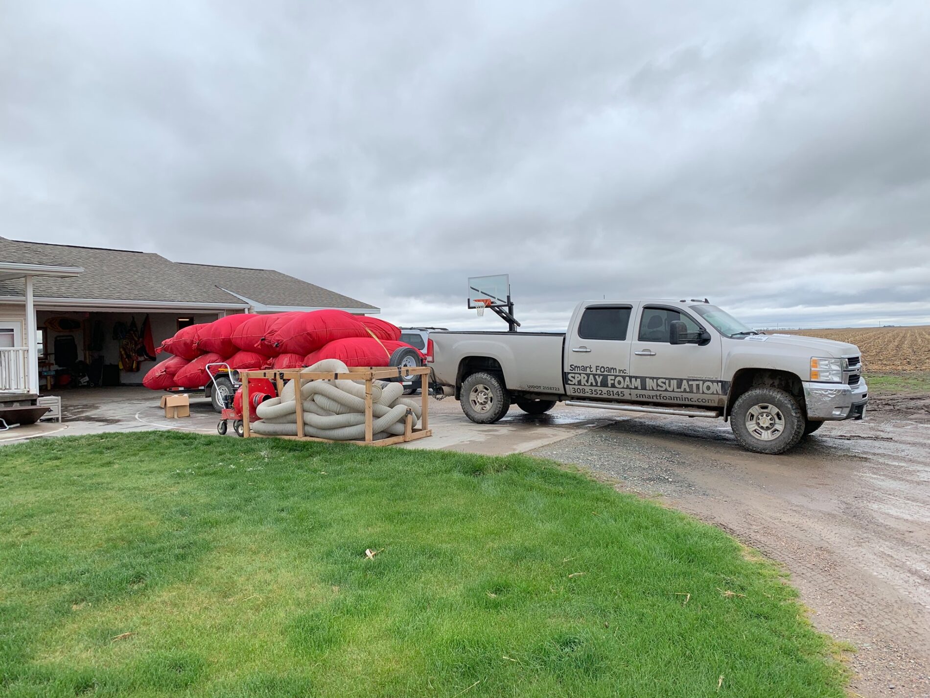 A truck labeled "Spray Foam Insulation" is parked in front of a house. Red tarps and coiled hoses are placed on the driveway. A person is seen handling equipment near the open garage.